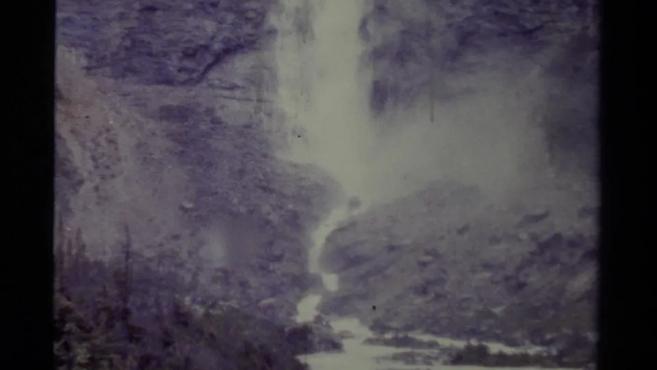 1979 Canadian 8mm frame showing a tall waterfall, mist, rocks, evergreens, and a stream in soft faded color.
