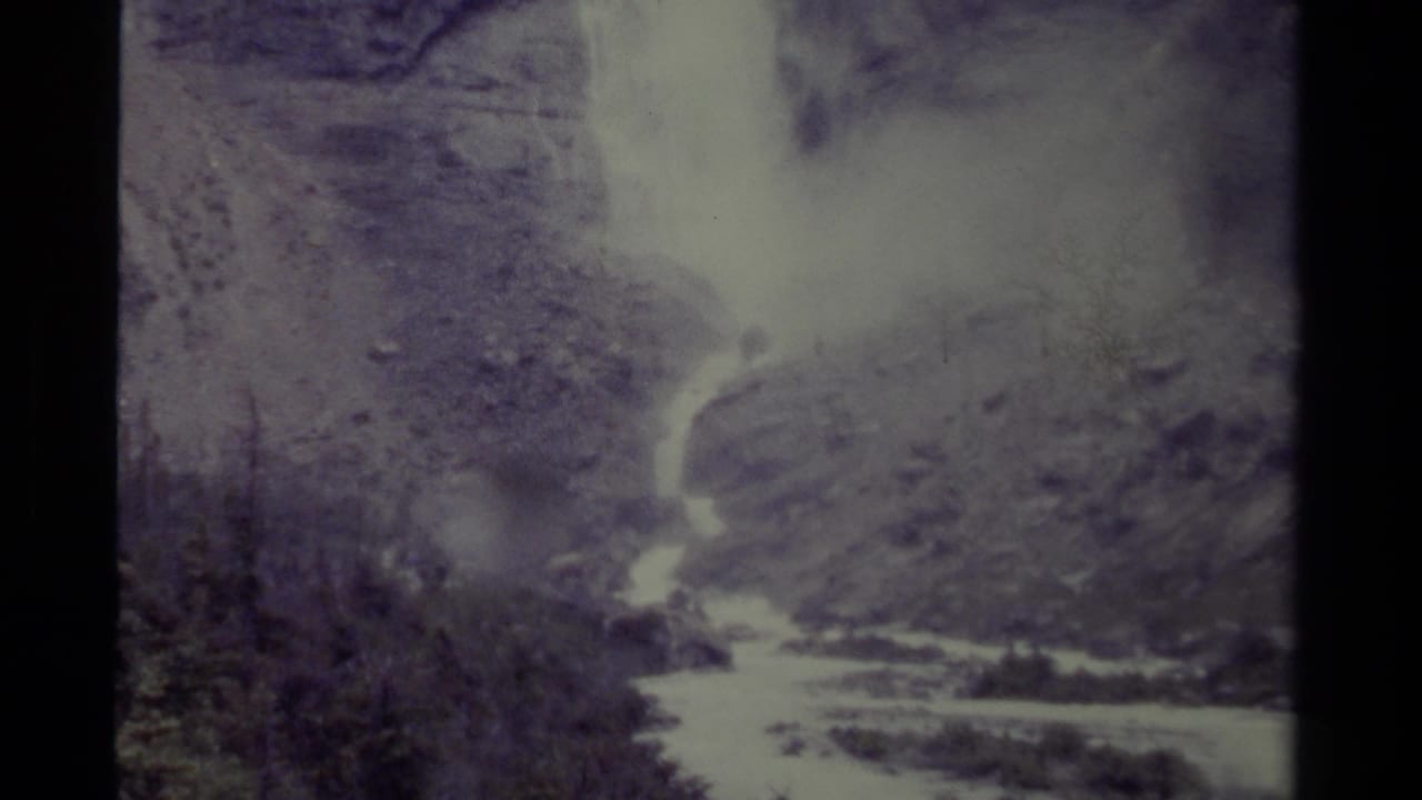 Misty waterfall and stream framed by rugged slopes and dark evergreen trees.