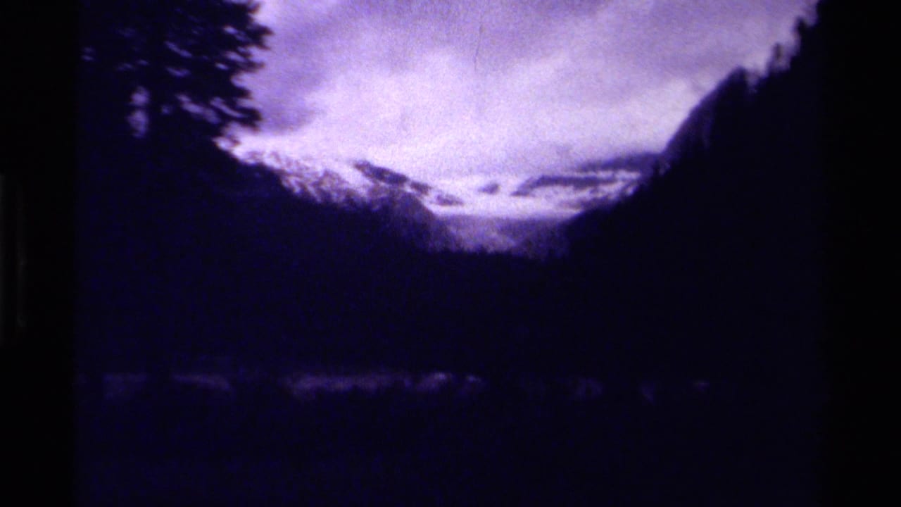 Snowy mountain pass seen between dark forested slopes under cloudy sky.
