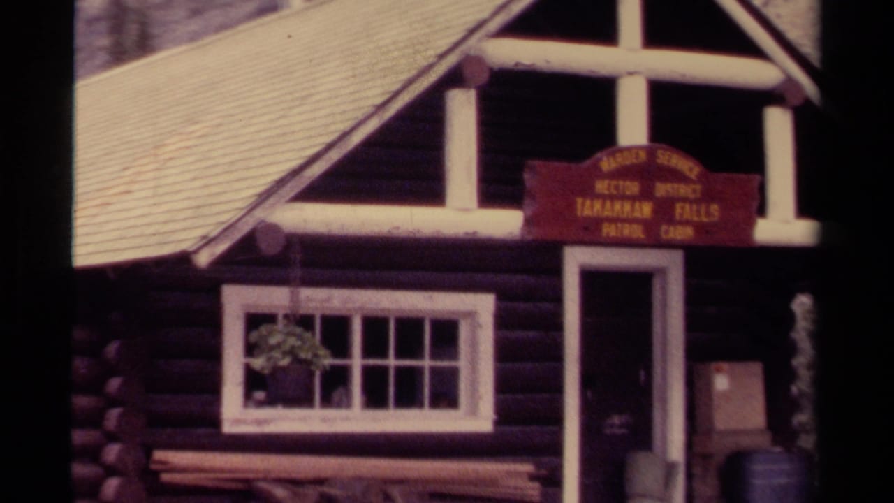 1979 Canada home-movie frame of the Takakkaw Falls Patrol Cabin facade with sign, open doorway, window, bench, and stacked firewood.