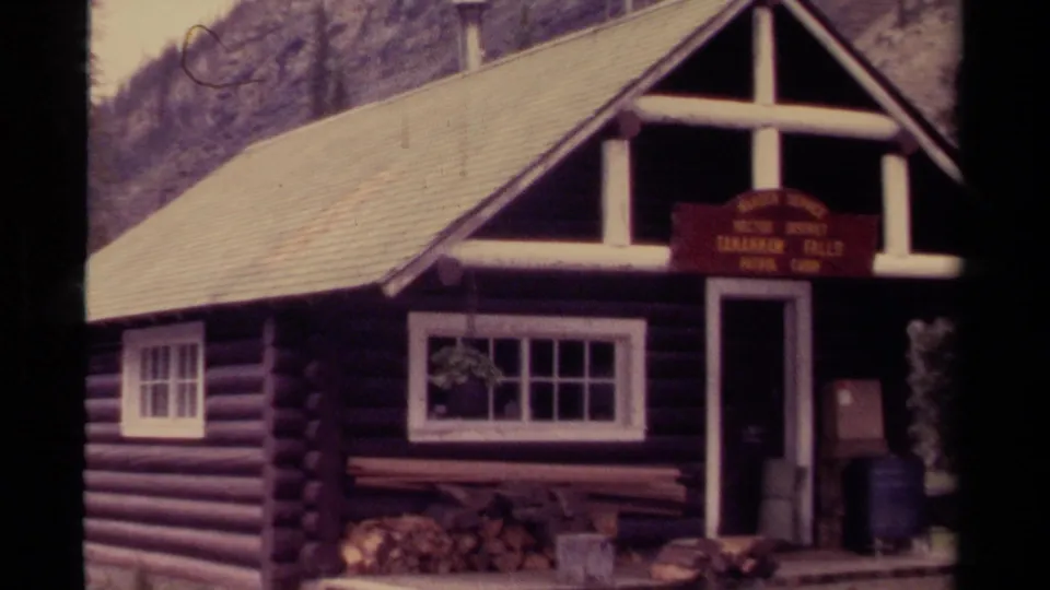 1979 8mm frame from Canada showing the patrol cabin, steep roof, white windows, red sign, and forested mountain backdrop.