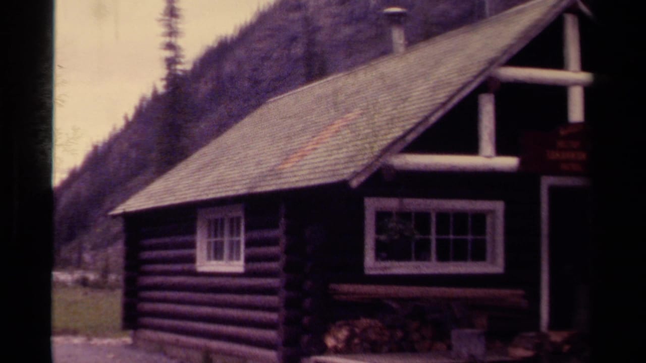 1979 8mm Canada footage showing a rustic log cabin near a river valley with evergreen-covered mountains behind it.