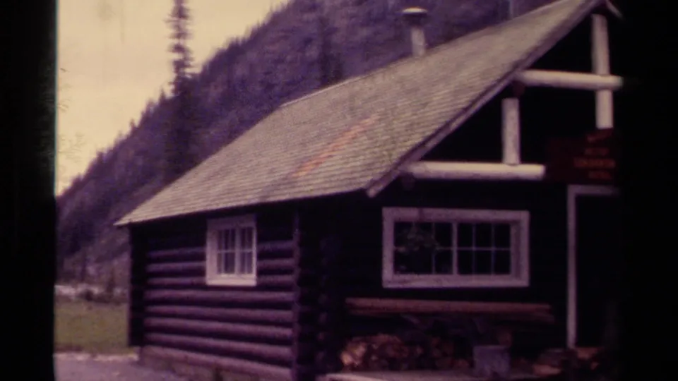 1979 8mm Canada footage showing a rustic log cabin near a river valley with evergreen-covered mountains behind it.