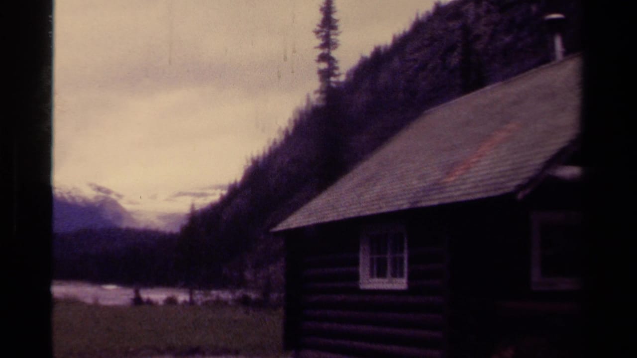 Visitor-shot 1979 Canada view of a cabin edge, river water, dark evergreen slopes, and distant snow-capped mountains.