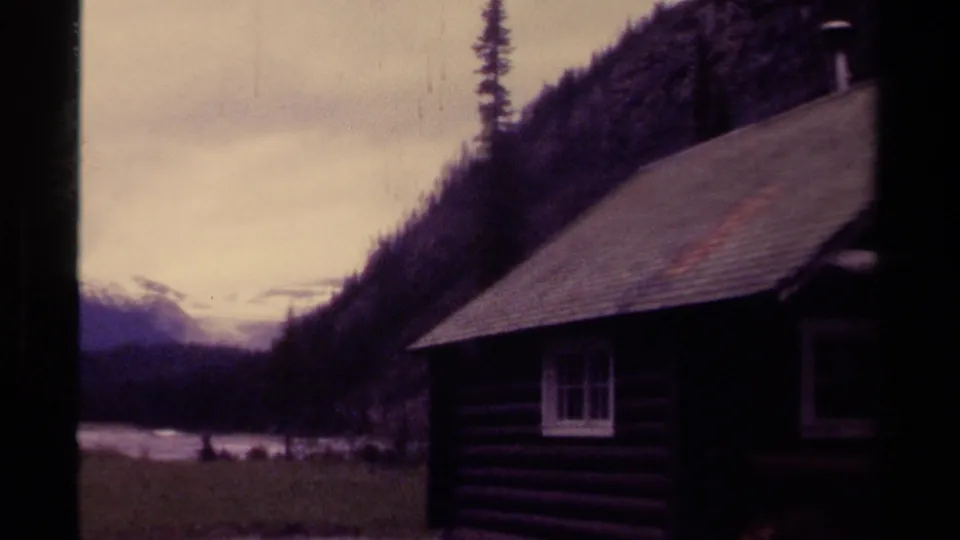 Visitor-shot 1979 Canada view of a cabin edge, river water, dark evergreen slopes, and distant snow-capped mountains.