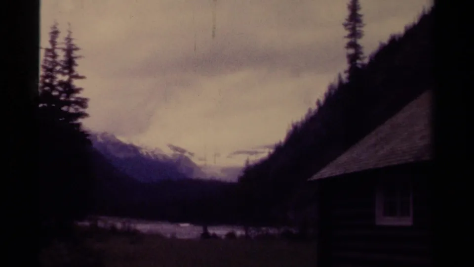 1979 Canadian home-movie frame looking past a cabin roof toward a river, evergreen trees, and snow-capped mountains.