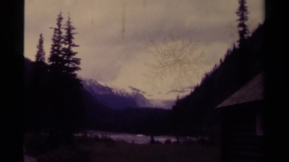 Faded 8mm Canada frame with evergreens, distant snowy mountains, cloudy sky, and the cabin roof cropped at right.