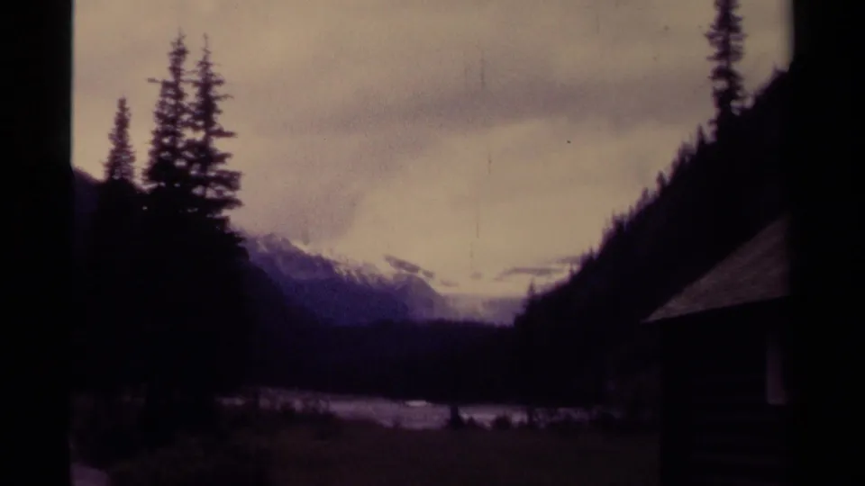 1979 Canada 8mm frame showing a shadowed mountain waterway, evergreen slopes, cloudy sky, and distant snow patches.