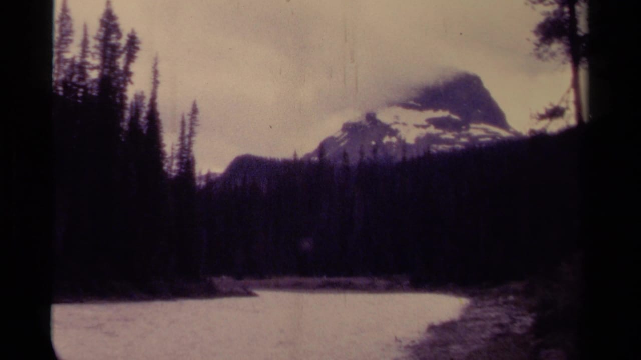 1979 Canada 8mm frame with a snow-capped peak, dense evergreen forest, still water, and a dark vertical foreground shape.
