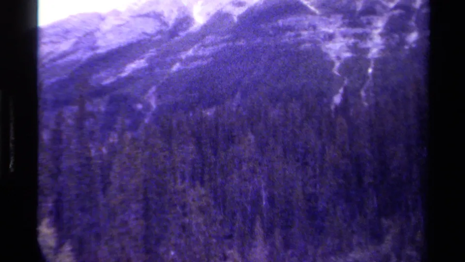 1979 Canadian 8mm frame showing dense evergreen forest below a snow-streaked mountain ridge, with dark vertical gate edges.