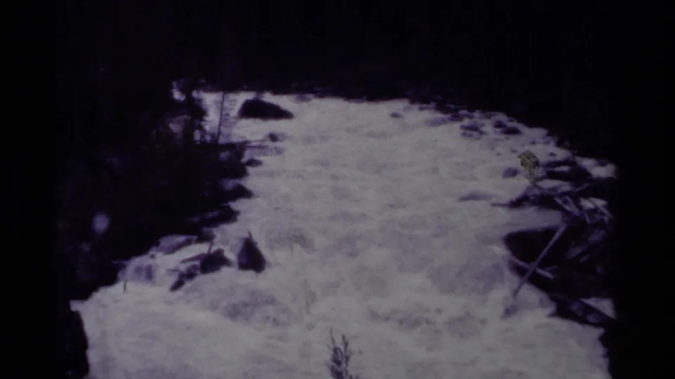 1979 Canadian 8mm frame showing foaming stream water rushing past rocks and fallen branches within a shadowed forest.
