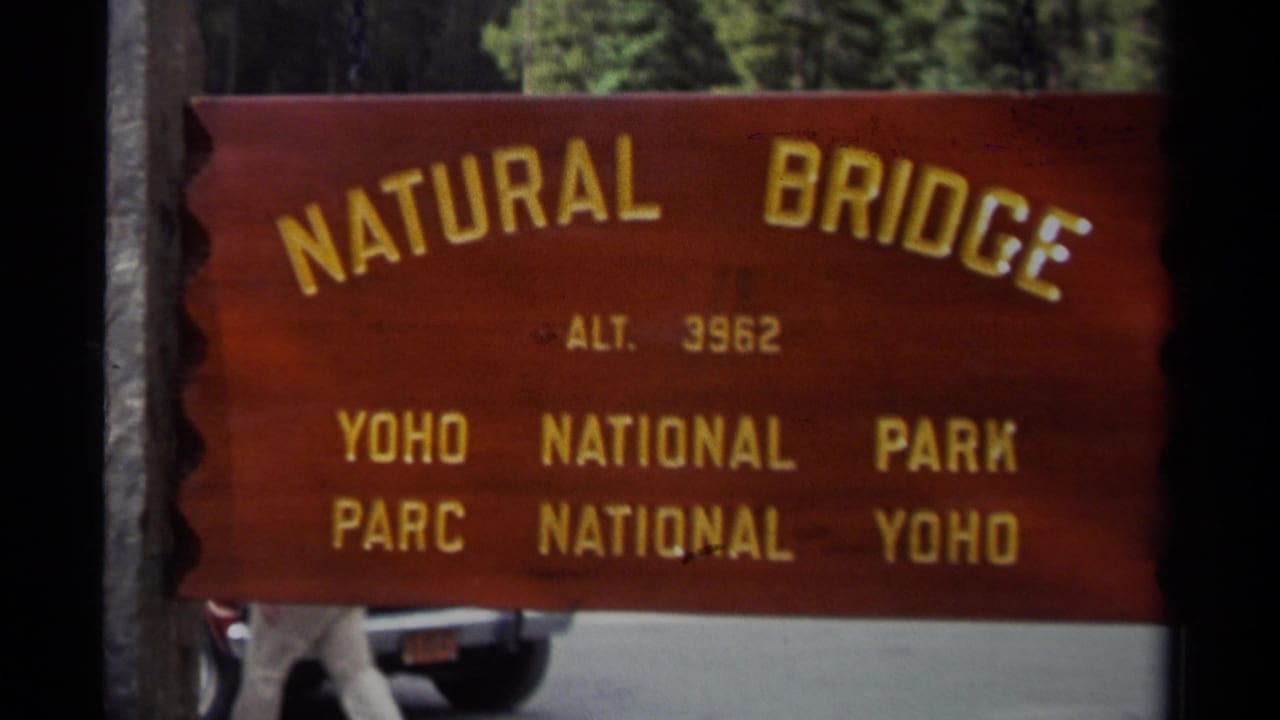 1979 Canadian home-movie frame of the Natural Bridge sign in Yoho National Park, with yellow lettering, trees, road, and a partial car.