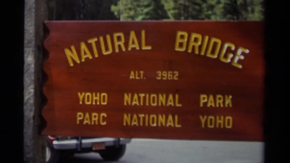 Visitor-shot 1979 frame of the Natural Bridge sign in Yoho National Park, with road and partial car visible below the sign.