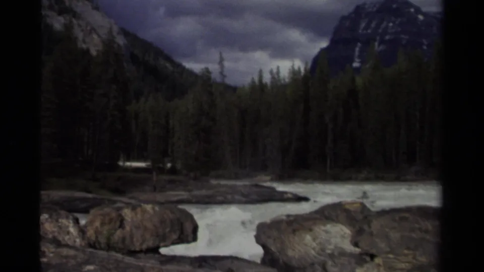 Yoho National Park river landscape with rushing water, boulders, conifer forest, steep mountains, and cloudy sky in 8mm footage.