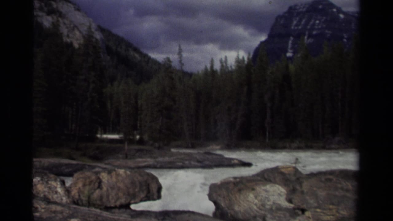 1979 8mm Canada footage showing pale river water, large rocks, dark evergreens, and mountain peaks under cloudy sky.