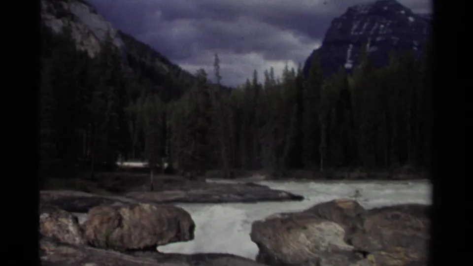 1979 8mm Canada footage showing pale river water, large rocks, dark evergreens, and mountain peaks under cloudy sky.