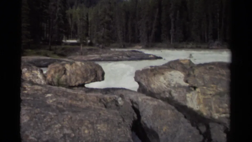 1979 home-movie frame of a mountain river flowing past large rocks, with dense evergreens beyond the water.
