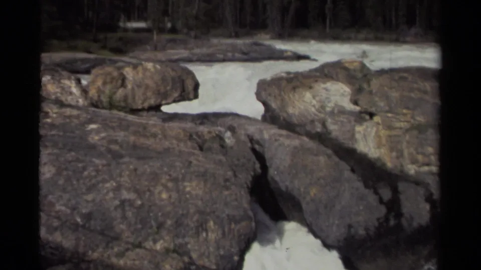 Final frame of the 1979 Canada 8mm clip showing water rushing between boulders below a dark evergreen forest.