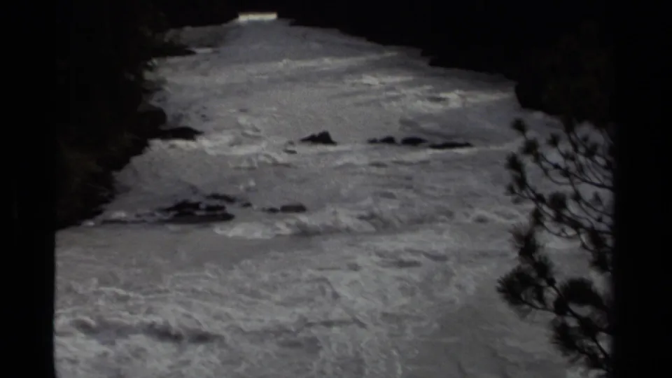 1979 Canadian 8mm frame showing turbulent river water bordered by dark evergreen forest and silhouetted trees.