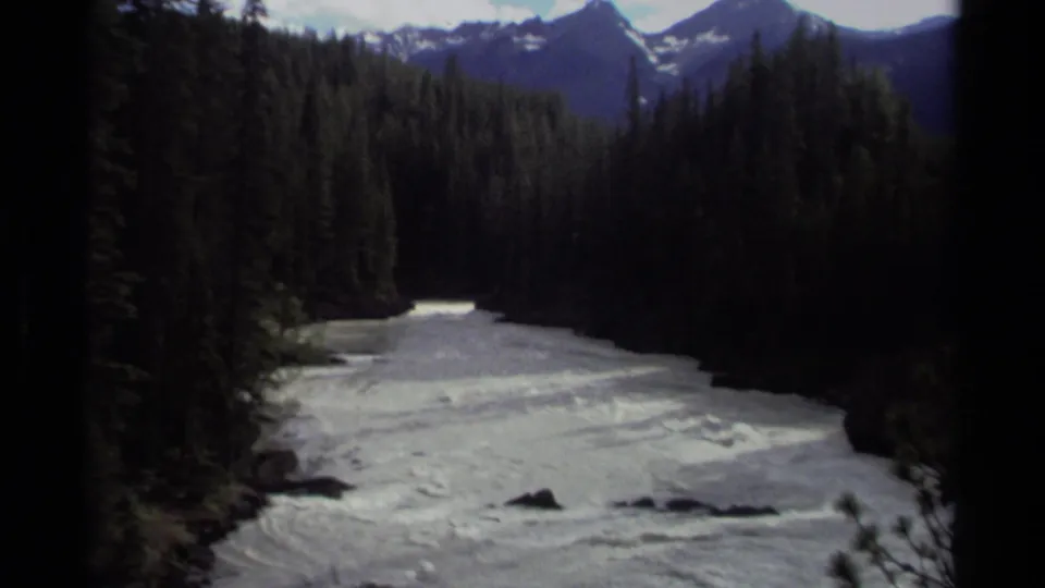 1979 Canadian 8mm frame showing a rushing forest river with distant snowy mountain peaks beyond the trees.