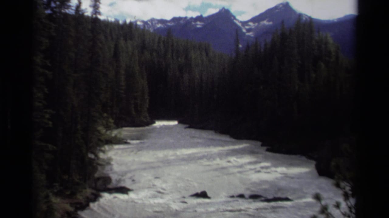 1979 Canadian 8mm frame showing a wide rushing river between dark evergreen forest and distant snow-capped peaks.
