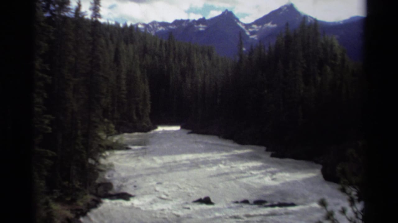 1979 Canadian 8mm view of a fast river framed by tall evergreens, with snowy mountain ridges in the background.