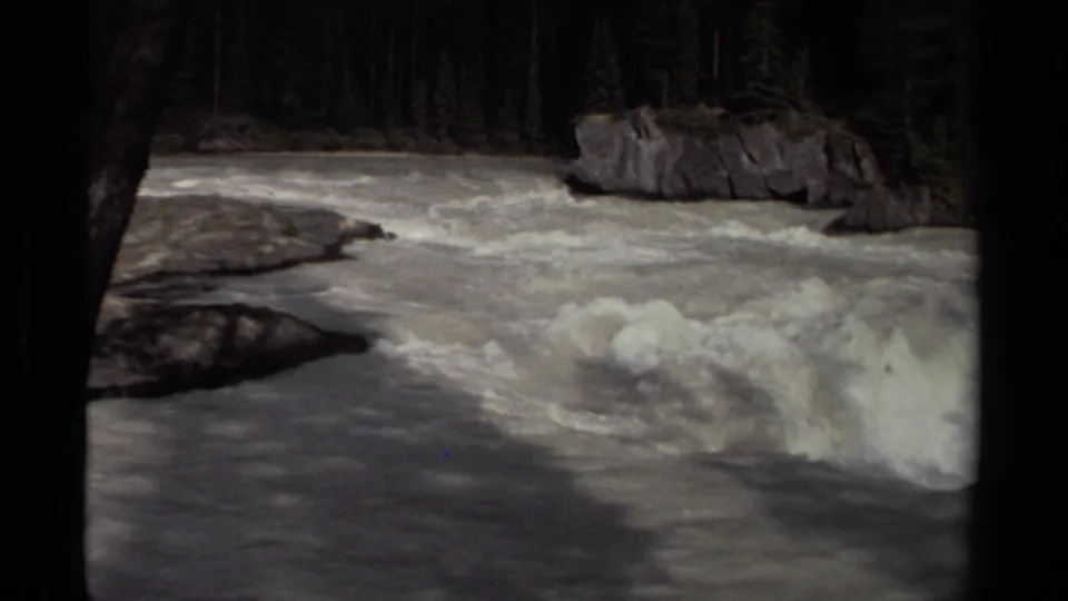 1979 Canadian 8mm frame with churning whitewater, exposed river rocks, and dark evergreen forest along the bank.