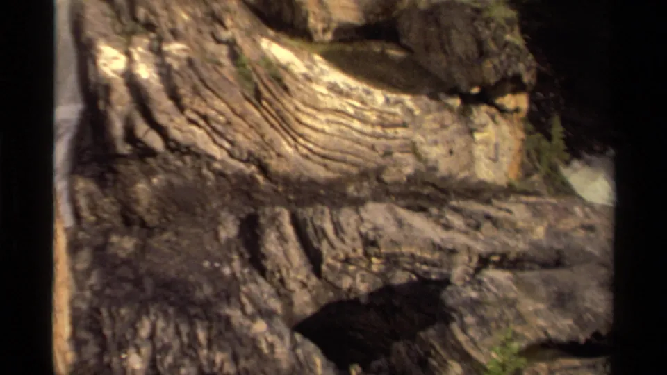 1979 Canada 8mm frame focused on rugged stone layers, dark recesses, and small green growth near waterfall spray.