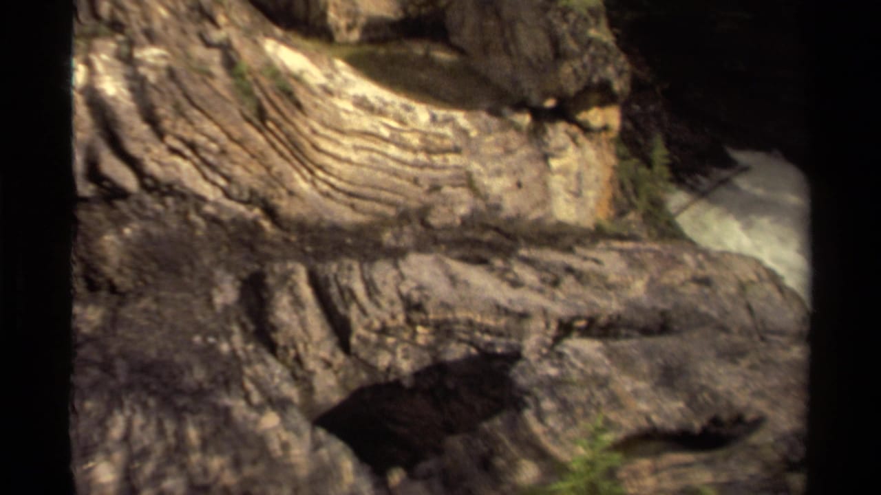 1979 Canadian 8mm frame showing striated riverside rock formations with fast white water passing at the edge of the view.
