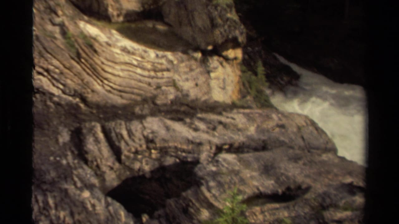 1979 Canadian 8mm frame of a rugged rock face and ledge with pale rushing water visible along the right side.