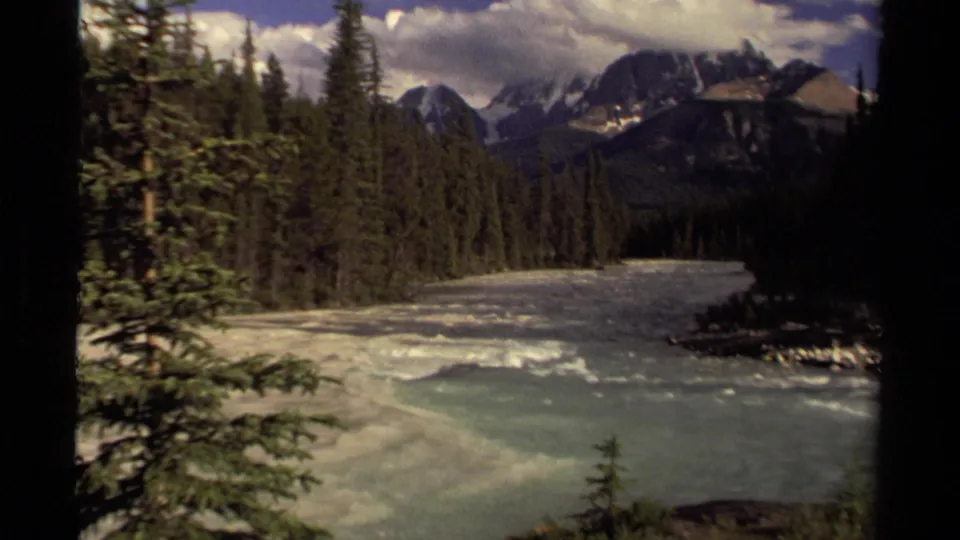 1979 Canadian 8mm frame showing a broad river, dense evergreen trees, distant dark mountains, snow patches, and cloudy sky.