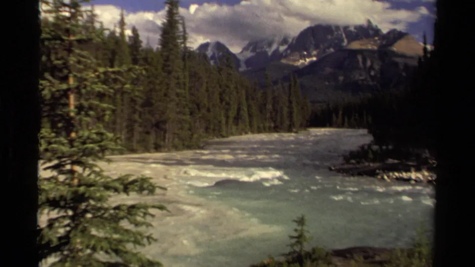 1979 Canadian 8mm frame with pale river water, pine forest, mountain ridges, small snow patches, and a partly cloudy sky.
