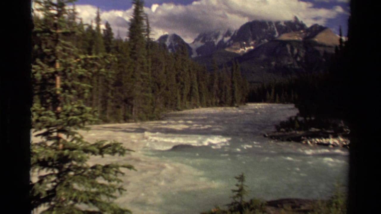 1979 Canadian home-movie frame showing fast river water through evergreen forest with distant mountains and clouded sky.