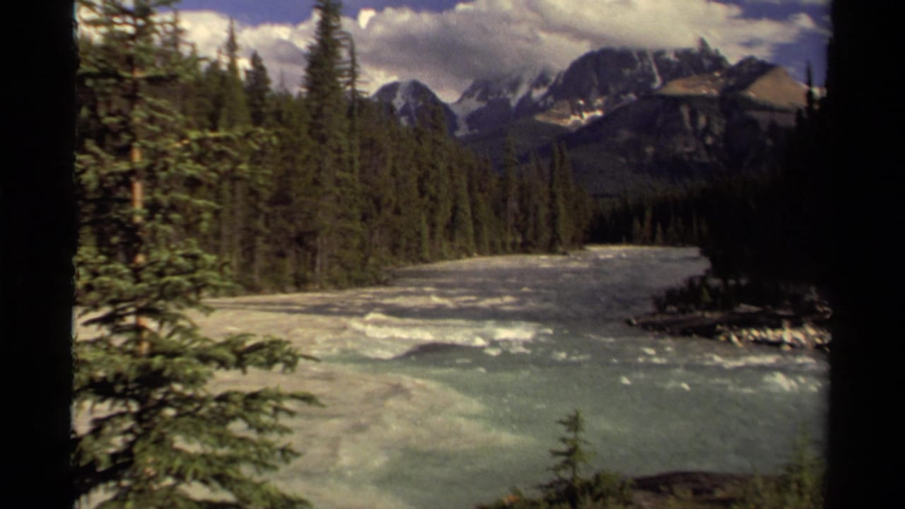 Wide view of churning river, evergreen forest, and mountains partly framed by dark film edges.