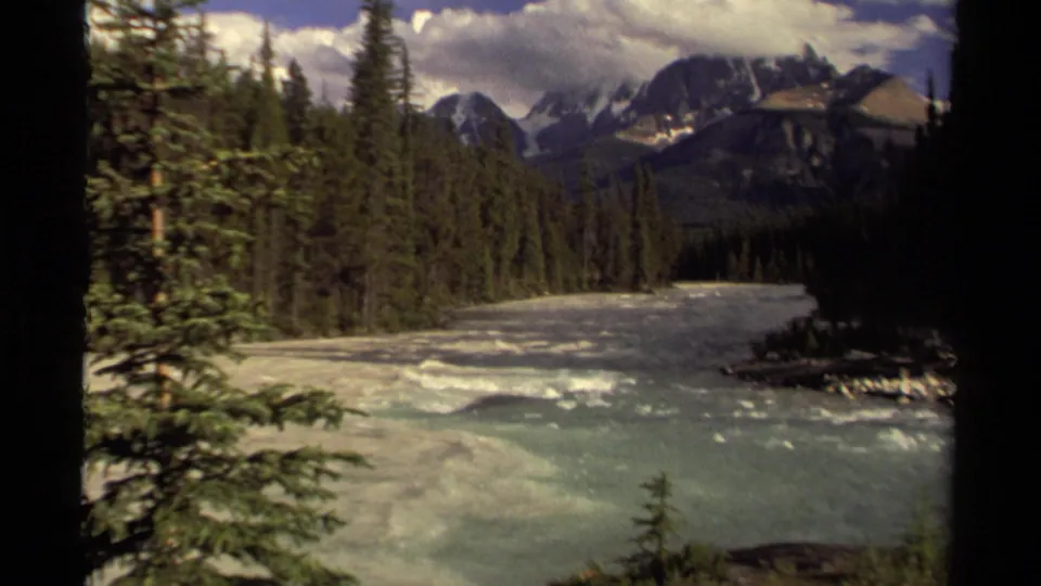 1979 Canadian wilderness footage frame with rolling water, conifers, distant mountain ridges, and heavy clouds.