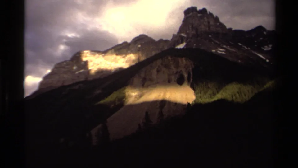 Vintage 8mm view of Canadian mountain peaks, with heavy clouds overhead and bands of sunlight across rocky slopes and snow.