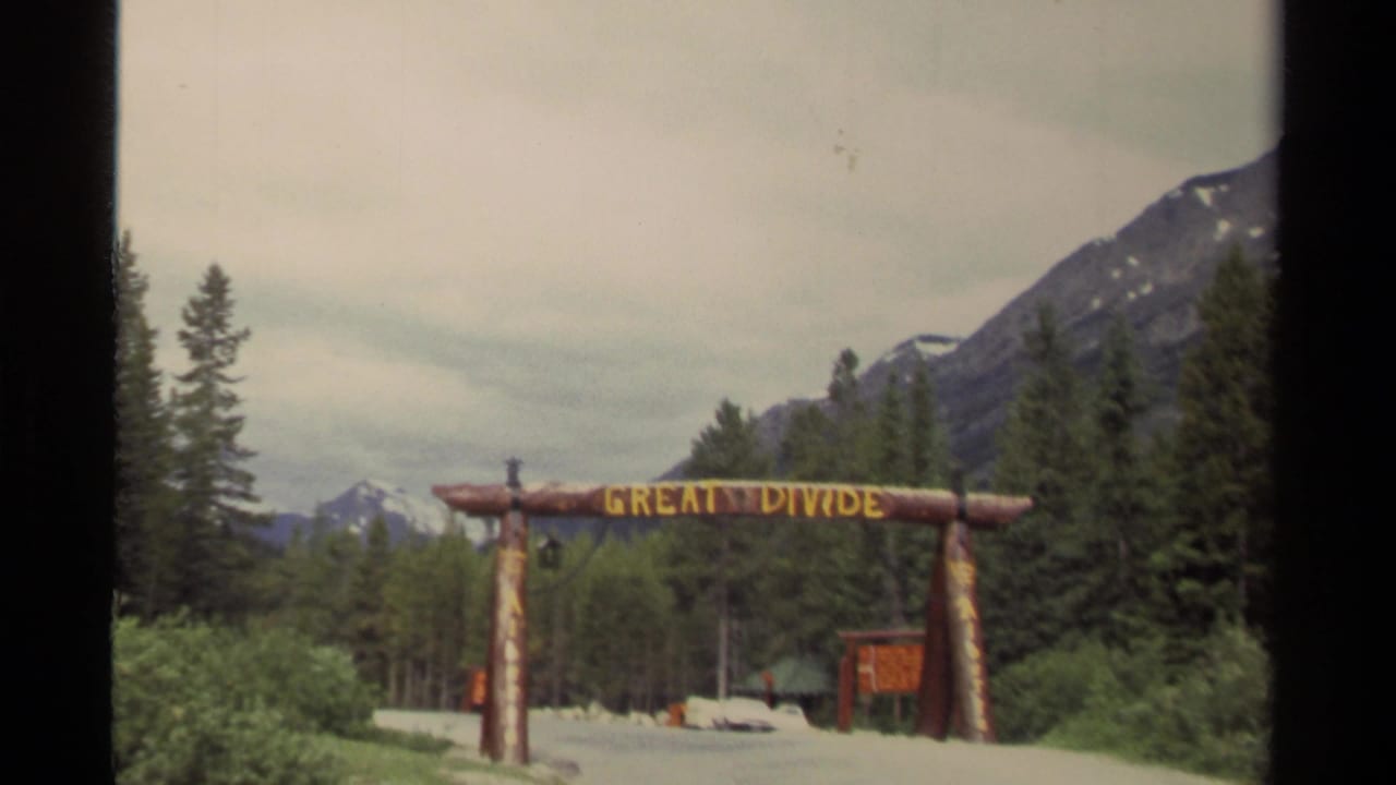 1979 Canada 8mm frame showing a rustic Great Divide log archway on a road bordered by evergreens and distant mountains.