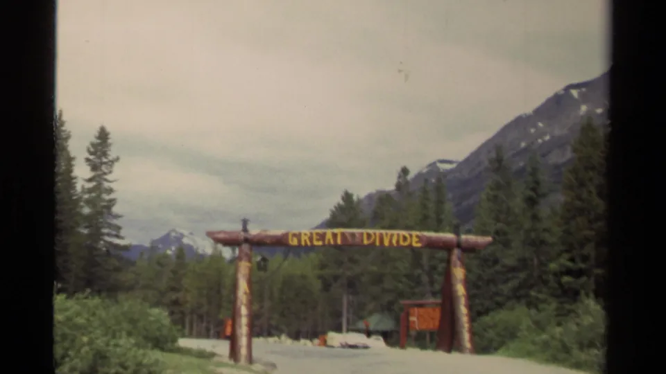 1979 Canada 8mm frame showing a rustic Great Divide log archway on a road bordered by evergreens and distant mountains.