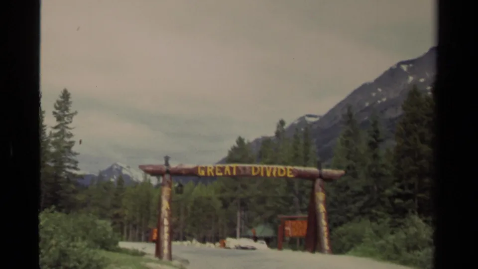 1979 Canada 8mm frame of the Great Divide archway seen from a visitor viewpoint with forest, cars, and mountain backdrop.
