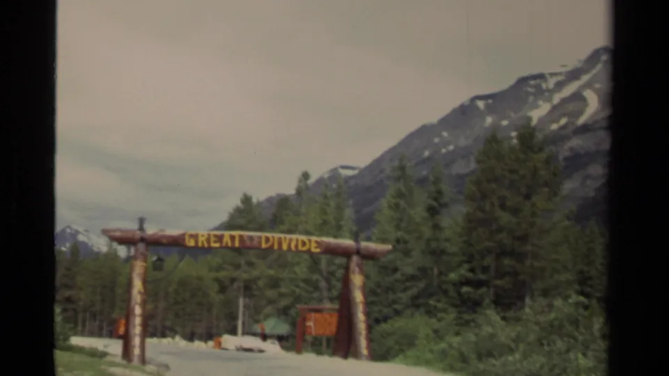 1979 Canada home-movie frame showing a rustic Great Divide log sign before forested mountain slopes.