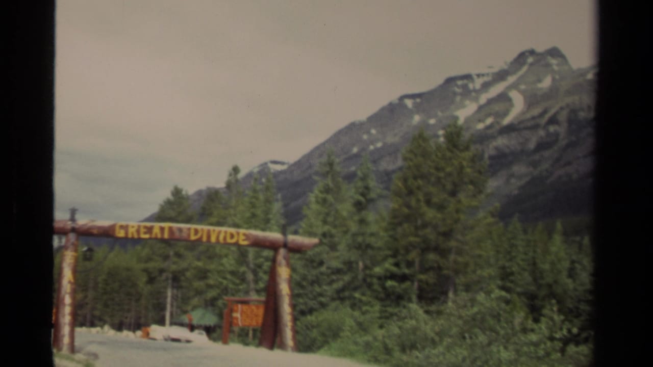 Vintage 8mm view of the Great Divide sign at a Canadian mountain roadside, framed by trees and peaks.