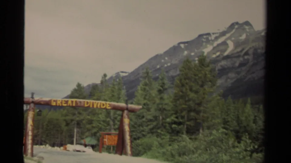 Vintage 8mm view of the Great Divide sign at a Canadian mountain roadside, framed by trees and peaks.