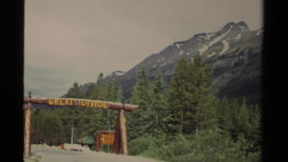 1979 Canada travel footage frame with the wooden Great Divide sign crossing the lower foreground below alpine scenery.