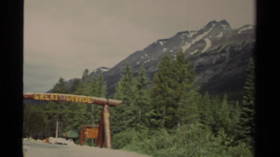 Canadian mountain-road home-movie frame where the Great Divide sign recedes as the camera passes dense evergreens.