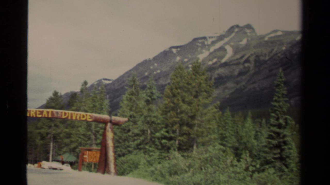 1979 Canada 8mm frame showing a Great Divide roadside sign, dense conifers, and snow-marked mountain ridges in the background.