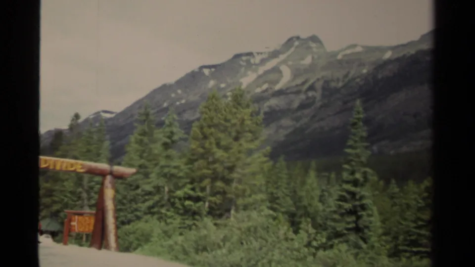 1979 Canada travel frame with the Great Divide sign at frame edge, conifer forest, and a broad rocky mountain slope under muted sky.