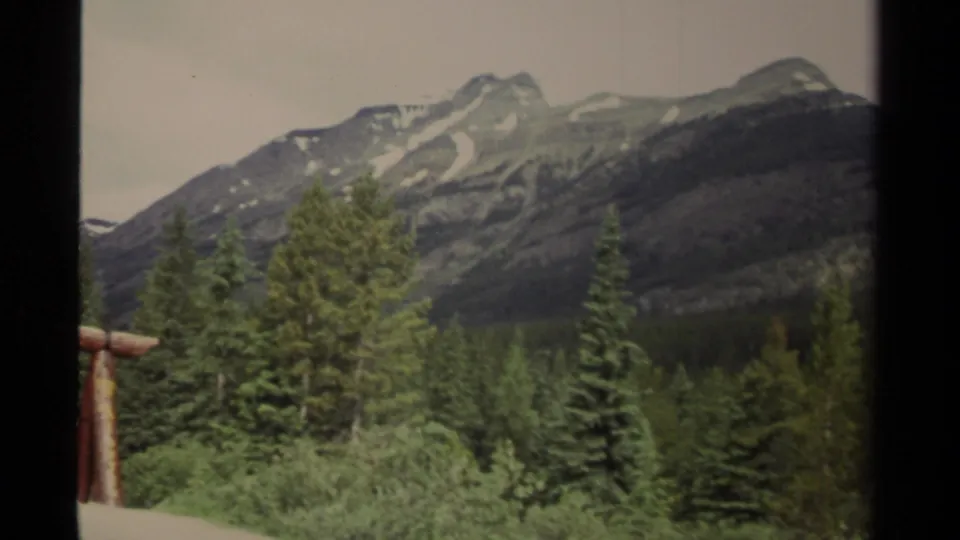 1979 Canada 8mm frame looking across roadside conifers toward a high mountain ridge, with only part of the signpost remaining at left.