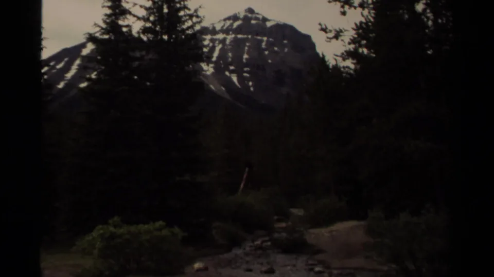 1979 Canada 8mm frame showing a shadowed mountain peak beyond tall trees, with a small creek or path running through the foreground.