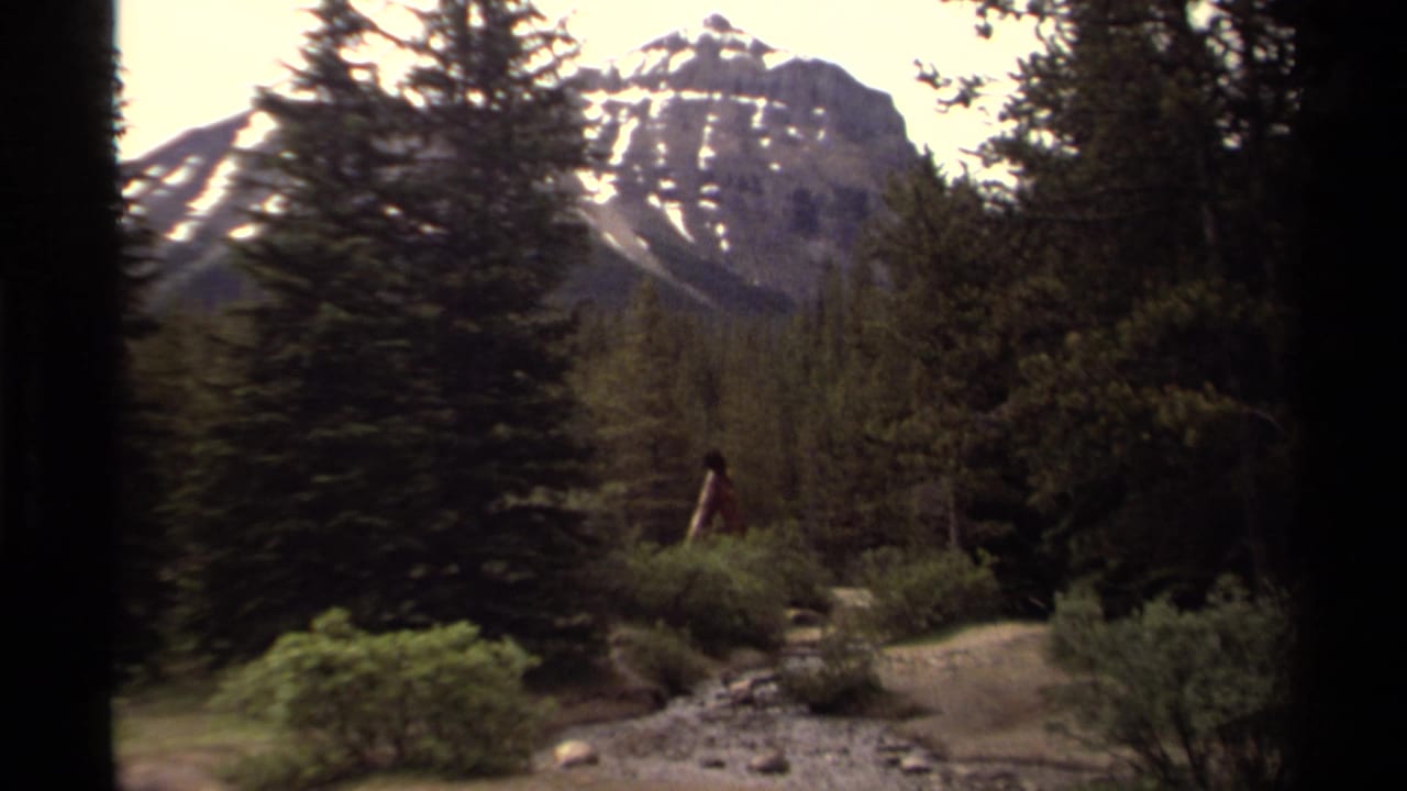 1979 Canadian 8mm frame with evergreens, a snow-streaked mountain, rocky ground, shrubs, and a distant walker in red.