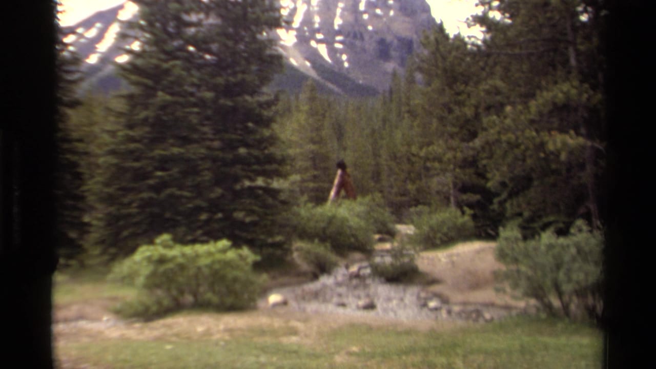 1979 Canadian mountain landscape with a distant walker, dense evergreens, low shrubs, rocky scree, and snow patches on the peak.